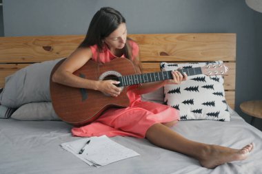 Pembe elbiseli esmer akustik gitarlı yatakta oturuyor ve yeni bir şarkı yazıyor. Müzisyen yeni bir şarkı bulur ve sözleri yazar..