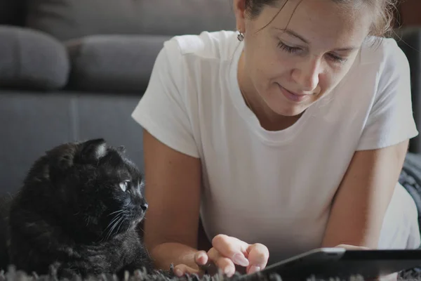 Gülümseyen yetişkin bir kadın elinde dijital tabletle evde yere uzanmış kitap okuyor. Kafkasyalı bir kız evcil hayvanıyla internet alışverişini yaparken bilgisayar uygulamaları kullanmaktan zevk alıyor.