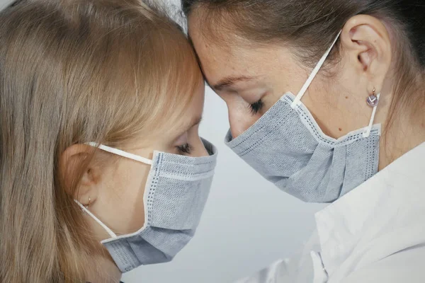 Close-up of female doctor wearing medical mask sitting face to face with little blonde girl in medical mask. The doctor calms the baby and hugs.
