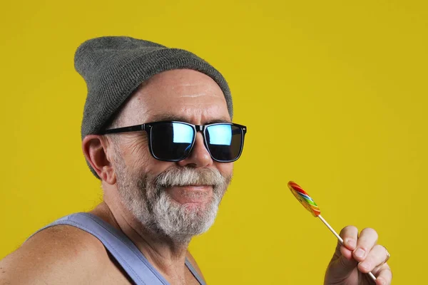サイドビューグレーの帽子とサングラスで高齢者のヒップスター男性のストック映像黄色の背景に対してカラフルな甘いロリポップをなめる タイムラプス — ストック写真