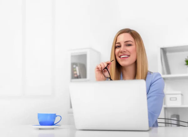 Attraente bionda in un abito blu è seduta alla scrivania dell'ufficio con un computer portatile e occhiali tra le mani . — Foto Stock