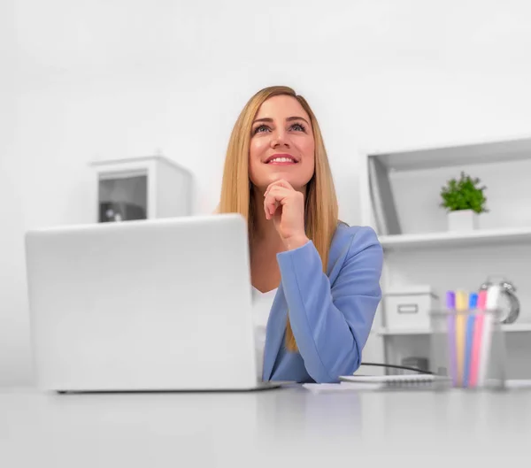 Attraktive blonde Frau, die verträumt nach oben schaut und mit Laptop an weißem Tisch im Büro sitzt. — Stockfoto