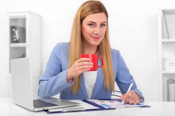 Bella bionda in un abito blu con una tazza rossa in mano al lavoro . — Foto Stock