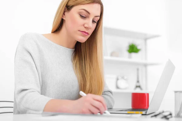 Une femme dans un bureau blanc est assise à une table et écrit. Une jeune femme utilisant un ordinateur portable remplit des documents sur le lieu de travail . — Photo