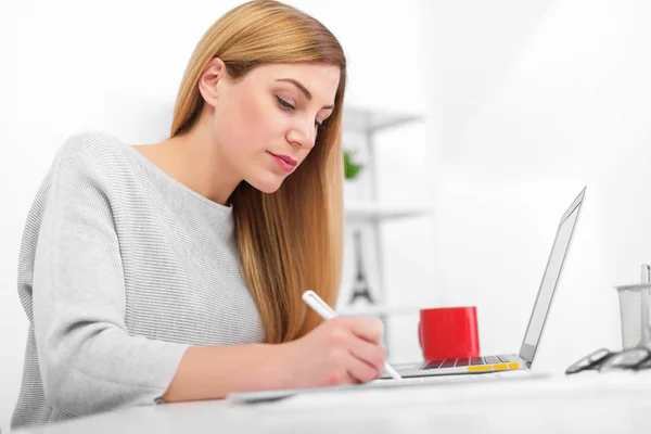 Una donna in un ufficio bianco è seduta a un tavolo a scrivere. Una giovane donna che utilizza un computer portatile riempie documenti sul posto di lavoro . — Foto Stock