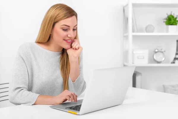 Přitažlivá blondýnka sedíte za stolem v kanceláři nebo doma při pohledu na obrazovku u notebooku. — Stock fotografie