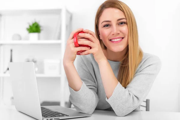 En söt ung kvinna håller en röd kopp sitter vid ett bord i en vit kontor. Fika. — Stockfoto