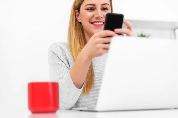 Jovem atraente com um smartphone na mão na mesa do escritório com um laptop . — Fotografia de Stock