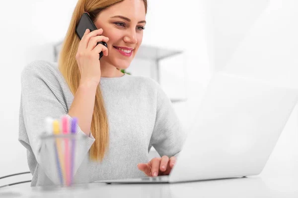 Attraktive junge Frau mit Smartphone in der Hand am Schreibtisch mit Laptop. — Stockfoto