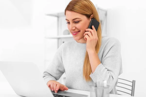 Atractiva joven con un teléfono inteligente en la mano en el escritorio de la oficina con un ordenador portátil . — Foto de Stock
