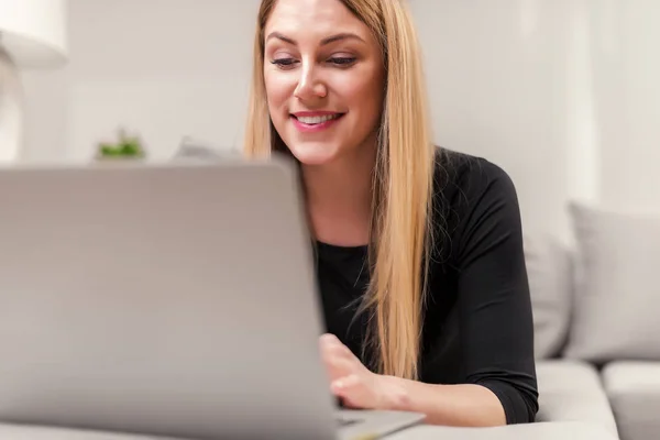 Aantrekkelijke vrouw met behulp van een laptop terwijl liggend op de Bank thuis. — Stockfoto