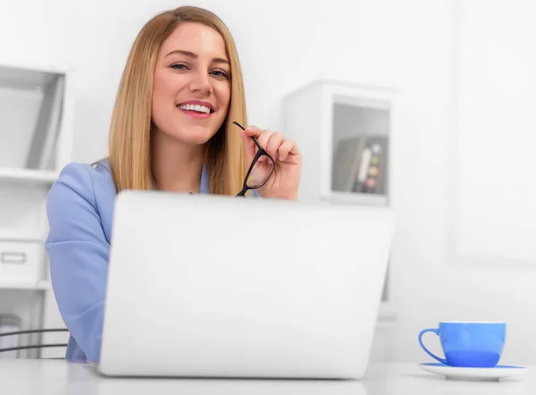 Jolie blonde en costume bleu est assise au bureau avec un ordinateur portable et des lunettes dans ses mains . — Photo