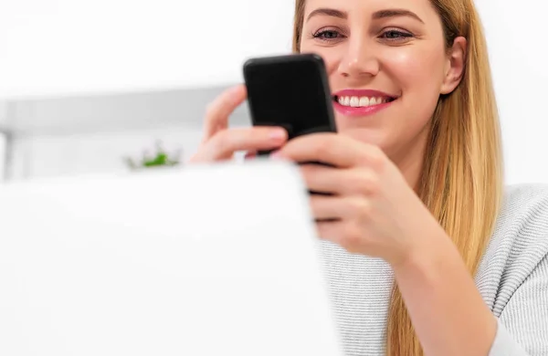 Attraktive junge Frau mit Smartphone in der Hand am Schreibtisch mit Laptop. — Stockfoto