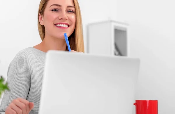 Uma loira muito bonita está sentada em uma mesa com um laptop e uma caneta na mão. Jovem mulher atraente no escritório ou no local de trabalho olhando para a câmera . — Fotografia de Stock