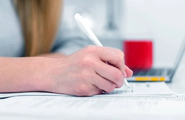 Una mujer escribe mientras está sentada en una mesa en una oficina o aula. La chica llena los documentos en el lugar de trabajo con una computadora portátil . —  Fotos de Stock