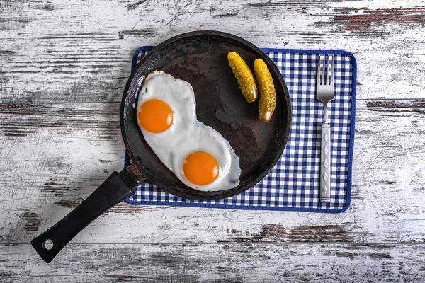 Fried eggs in a frying pan. Rye toast and pickled cucumber next to fried eggs.