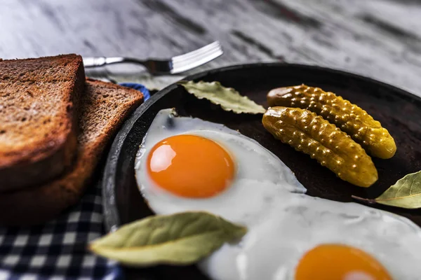 Fried eggs in a frying pan. Rye toast and pickled cucumber next to fried eggs. — Stock Photo, Image