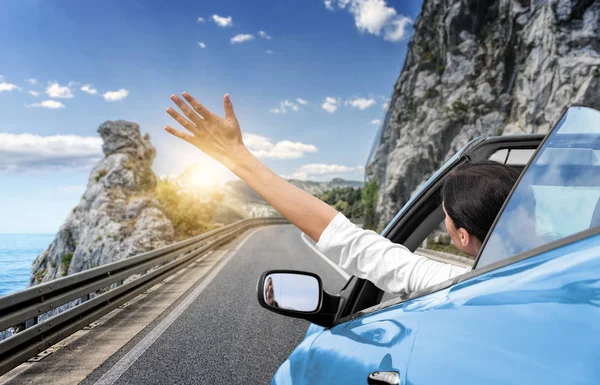 Una mujer joven está conduciendo en coche hacia el mar y agitando su mano desde un coche descapotable azul. Vacaciones en la costa del mar . —  Fotos de Stock