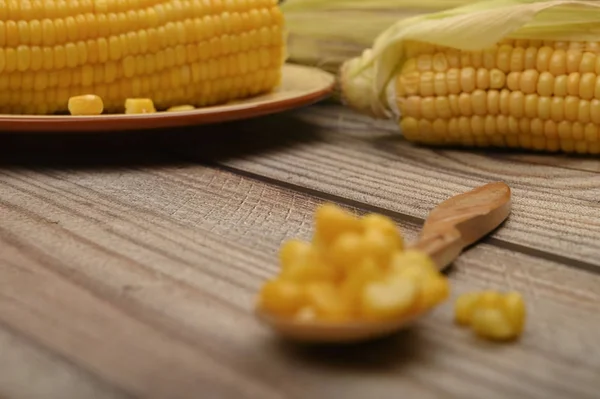 Grains of sweet corn on a wooden spoon on a wooden table, in the background boiled corn and untreated ears of corn. Fitness diet. Healthy diet. For a sweet treat. Close up. Selective focus. — Stock Photo, Image