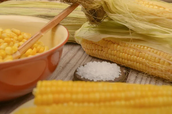 Grains of sweet corn in a plate with a wooden spoon, coarse salt and ears of corn on the table. Healthy diet. Fitness diet. For a sweet treat. Close up.