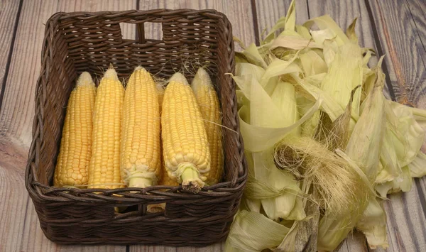 Peeled ears of corn in a wicker basket and corn leaves on a wooden table. Autumn harvest. Fitness diet. Healthy diet. For a sweet treat. — Stock Photo, Image