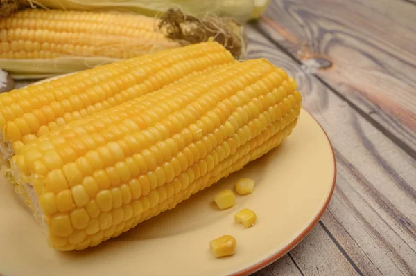 Два вареных колоска кукурузы на тарелке и сырые колосья кукурузы на деревянном столе. Здоровое питание. Фитнес-диета. За сладкое угощение. Закрыть . — стоковое фото