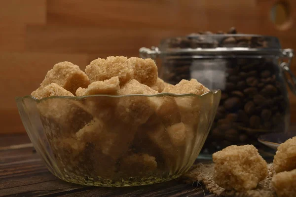 Pieces of brown sugar in a sugar bowl and coffee beans in a glass jar on a wooden background. Close up.