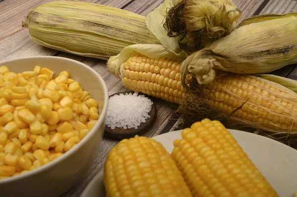 Два вареных колоска кукурузы, зерна кукурузы, грубая соль и сырые колосья кукурузы на деревянном столе. Фитнес-диета. Здоровое питание. За сладкое угощение. Закрыть . — стоковое фото
