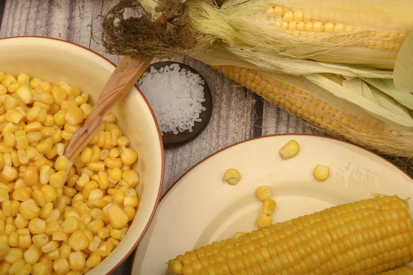 Зерна сладкой кукурузы в тарелке с деревянной ложкой, грубой солью и колосьями кукурузы на столе. Здоровое питание. Фитнес-диета. За сладкое угощение. Закрыть . — стоковое фото