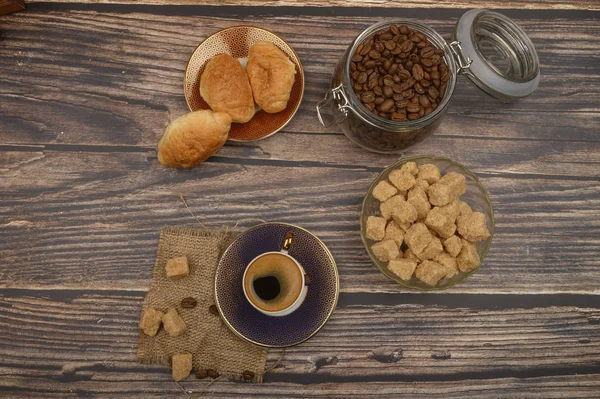 Eine Tasse Kaffee, braune Zuckerstücke in einer Zuckerdose, Kaffeebohnen im Glas und Croissants auf einem hölzernen Hintergrund. — Stockfoto