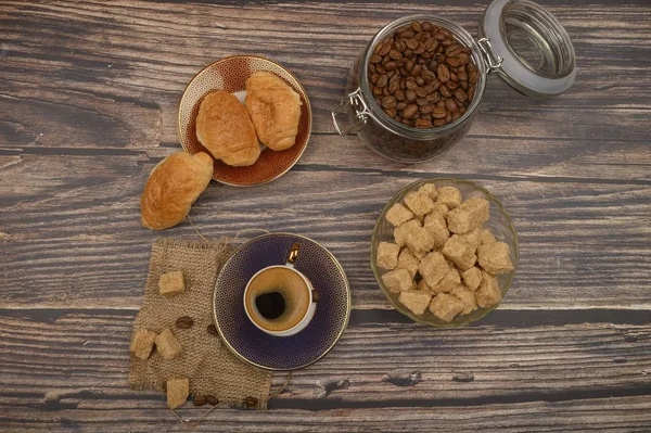 Una taza de café, trozos de azúcar morena en un tazón de azúcar, granos de café en un frasco de vidrio y croissants sobre un fondo de madera . — Foto de Stock