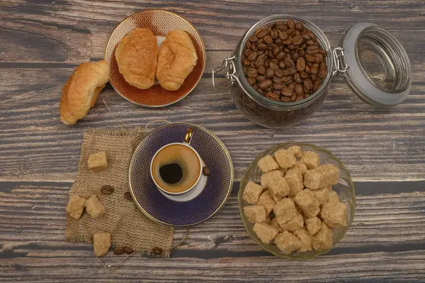 Eine Tasse Kaffee, braune Zuckerstücke in einer Zuckerdose, Kaffeebohnen im Glas und Croissants auf einem hölzernen Hintergrund. — Stockfoto