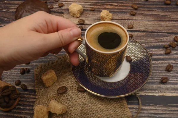 Die Hand des Mädchens hält eine Tasse Kaffee über einen Holztisch mit braunem Zucker und Kaffeebohnen. — Stockfoto