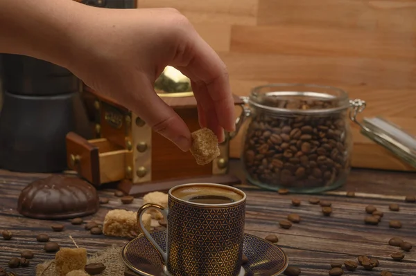 Hand Mädchen legt ein Stück braunen Zucker in eine Tasse Kaffee, Kaffeebohnen, braunen Zucker, Kaffeemühle auf einem hölzernen Hintergrund. — Stockfoto