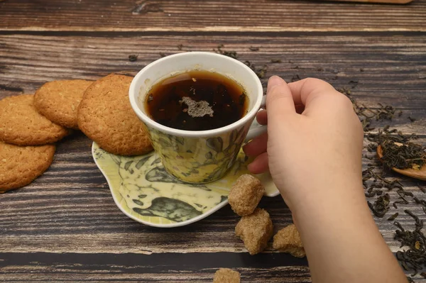 A mão da menina segura uma caneca de chá preto, biscoitos de aveia, folhas de chá, açúcar mascavo em um fundo de madeira. Fechar . — Fotografia de Stock