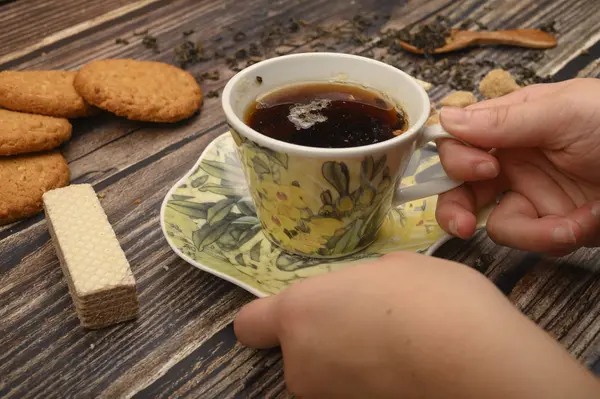 La mano de la niña sostiene una taza de té negro, galletas de avena, gofres, hojas de té, azúcar morena sobre un fondo de madera. De cerca. . — Foto de Stock