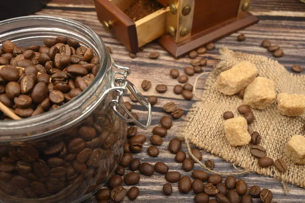 Cuillère en bois dans un bocal en verre avec grains de café, moulin à café, morceaux de sucre brun sur un fond en bois. Gros plan . — Photo