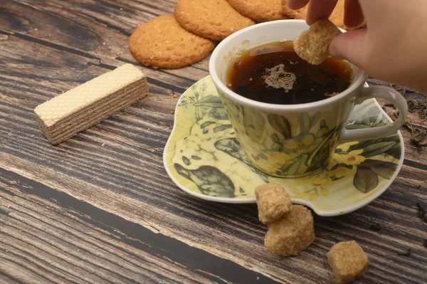 La mano de la niña pone un trozo de azúcar morena en una taza de té negro, galletas de avena, hojas de té, gofres, azúcar morena sobre un fondo de madera. De cerca. . — Foto de Stock