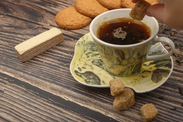 La main de la fille met un morceau de sucre brun dans une tasse de thé noir, des biscuits à l'avoine, des feuilles de thé, des gaufres, de la cassonade sur un fond en bois. Gros plan . — Photo