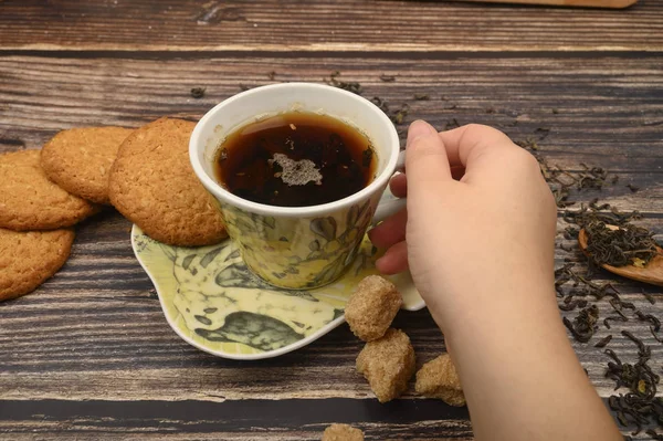 La mano de la niña sostiene una taza de té negro, galletas de avena, hojas de té, azúcar morena sobre un fondo de madera. De cerca. . — Foto de Stock