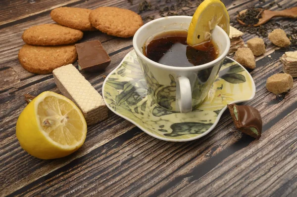 Eine Tasse schwarzen Tee, Zitronenscheiben, braune Zuckerscheiben, Haferflockenkekse, Waffeln auf einem hölzernen Hintergrund. Nahaufnahme. — Stockfoto