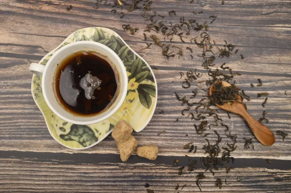 Une tasse de thé noir, des feuilles de thé dans une cuillère en bois, des morceaux de sucre brun sur un fond en bois. Gros plan . — Photo