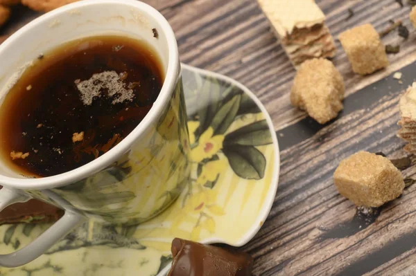 A Cup of black tea, tea leaves, pieces of brown sugar, oatmeal cookies, waffles, pieces of chocolate on a wooden background. Close up. — Stock Photo, Image