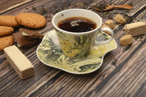 Eine Tasse schwarzen Tee, Teeblätter, braunen Zucker, Haferflockenkekse, Waffeln, Schokoladenstücke auf einem hölzernen Hintergrund. Nahaufnahme. — Stockfoto