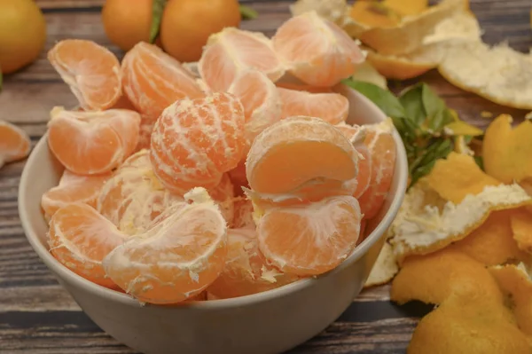 Peeled tangerines in a ceramic dish, tangerines on a twig with green leaves, tangerine peel on a wooden background. Autumn harvest. Close up. — Stock Photo, Image