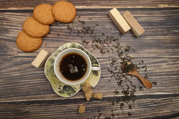 Una taza de té negro, hojas de té, trozos de azúcar morena, galletas de avena, gofres sobre un fondo de madera. De cerca. . — Foto de Stock