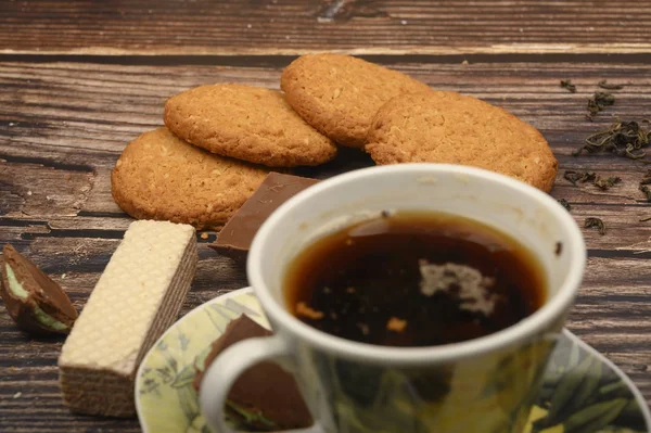 Uma xícara de chá preto, folhas de chá, pedaços de açúcar mascavo, biscoitos de aveia, waffles, pedaços de chocolate em um fundo de madeira. Fechar . — Fotografia de Stock