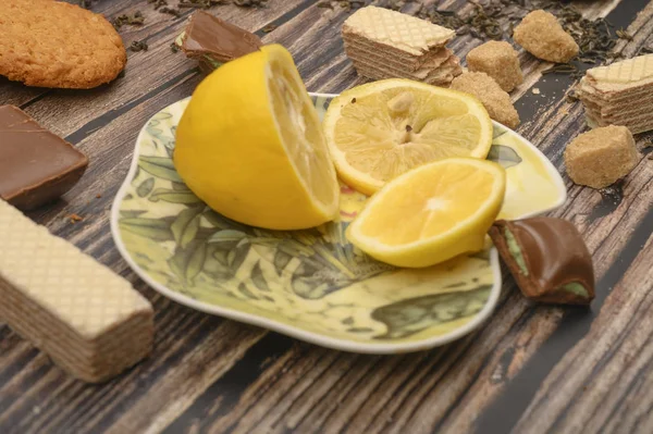 Sliced lemon on a saucer, pieces of brown sugar, oatmeal cookies, waffles, pieces of chocolate on a wooden background. Close up. — Stock Photo, Image