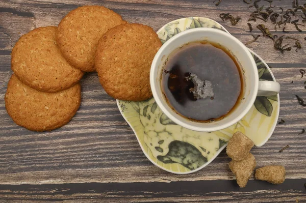 Una taza de té negro, hojas de té, trozos de azúcar morena, galletas de avena sobre un fondo de madera. De cerca. . — Foto de Stock