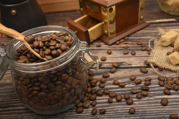 Cuillère en bois dans un bocal en verre avec grains de café, moulin à café, morceaux de sucre brun sur un fond en bois. Gros plan . — Photo